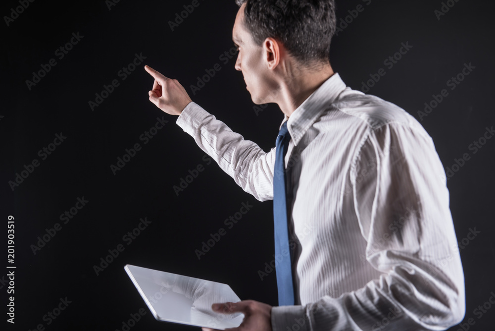 Digital screen. Intelligent nice adult mans standing with a tablet and pointing with his hand while looking at the digital screen Digital screen. Intelligent nice adult mans standing with a tablet and pointing with his hand while looking at the digital screen