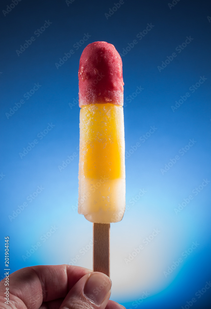 gefrorenes fruchtiges tropisches Eis am Stiel rot gelb weiß in der Hand ...