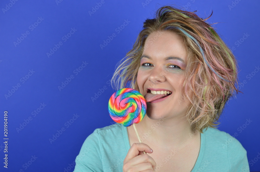 portrait of a young blond woman with colourful hair licking at a big lollipop and looking into the camera