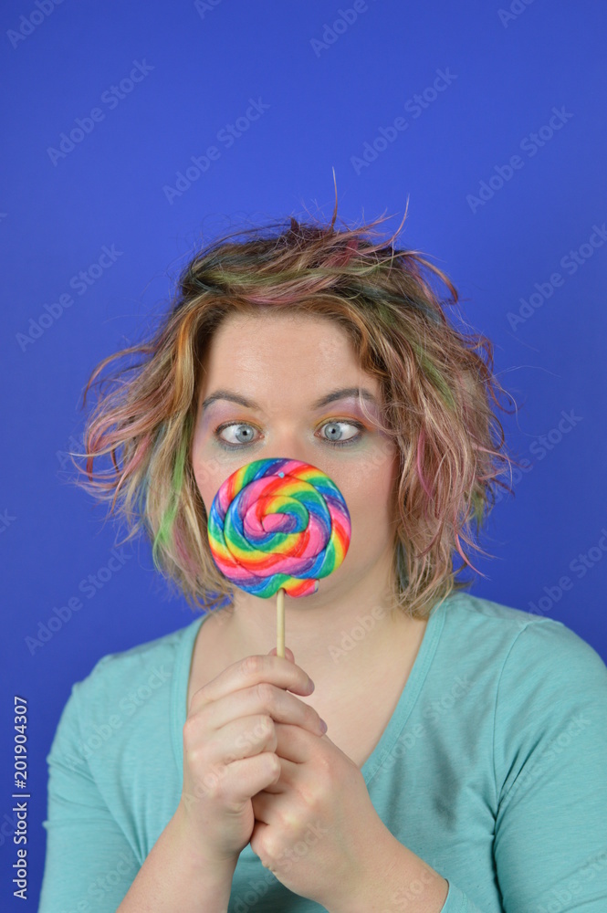 portrait of a young blond woman with colourful streaks in the hair and a big sweet multicolored lollipop in her hands squinting and having fun