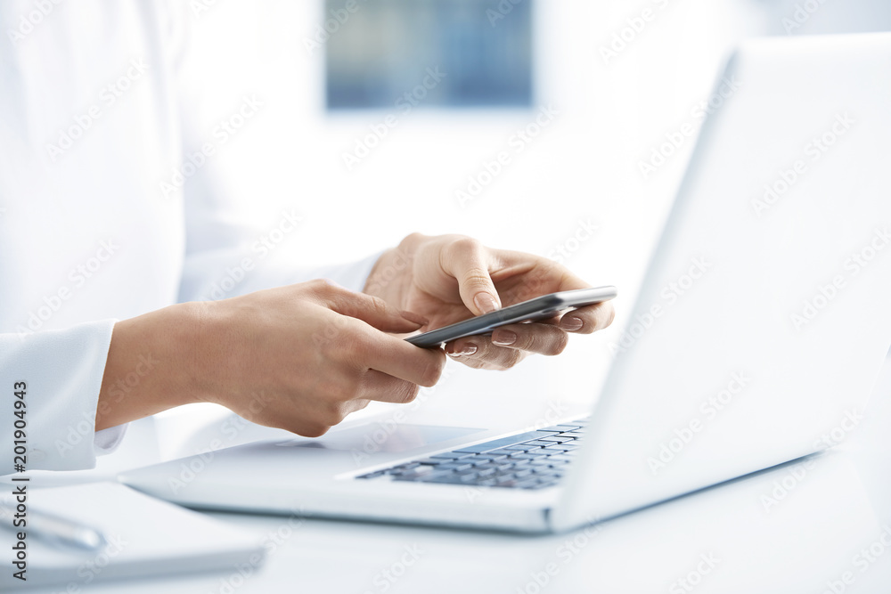 Fototapeta premium Woman using her mobile phone and send message. Close-up shot of businesswoman holding cellphone in her hands and text messaging while sitting at office desk in front of laptop. 