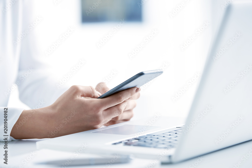 Fototapeta premium Woman using her mobile phone and send message. Close-up shot of businesswoman holding cellphone in her hands and text messaging while sitting at office desk in front of laptop. 