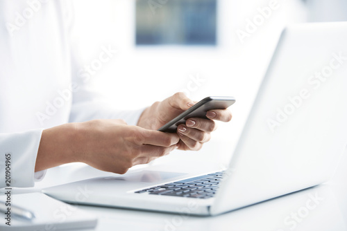 Woman using her mobile phone and send message. Close-up shot of businesswoman holding cellphone in her hands and text messaging while sitting at office desk in front of laptop. 