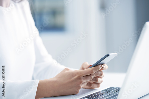 Woman using her mobile phone and send message. Close-up shot of businesswoman holding cellphone in her hands and text messaging while sitting at office desk in front of laptop. 