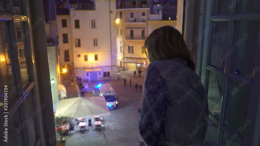 A night incident in the city. Ambulance car on the street. Excited Young woman looks out the window of her house. First aid, life insurance, terrorist threat. Home quarantine coronavirus Italy A night incident in the city. Ambulance car on the street. Excited Young woman looks out the window of her house. First aid, life insurance, terrorist threat. Home quarantine coronavirus Italy
