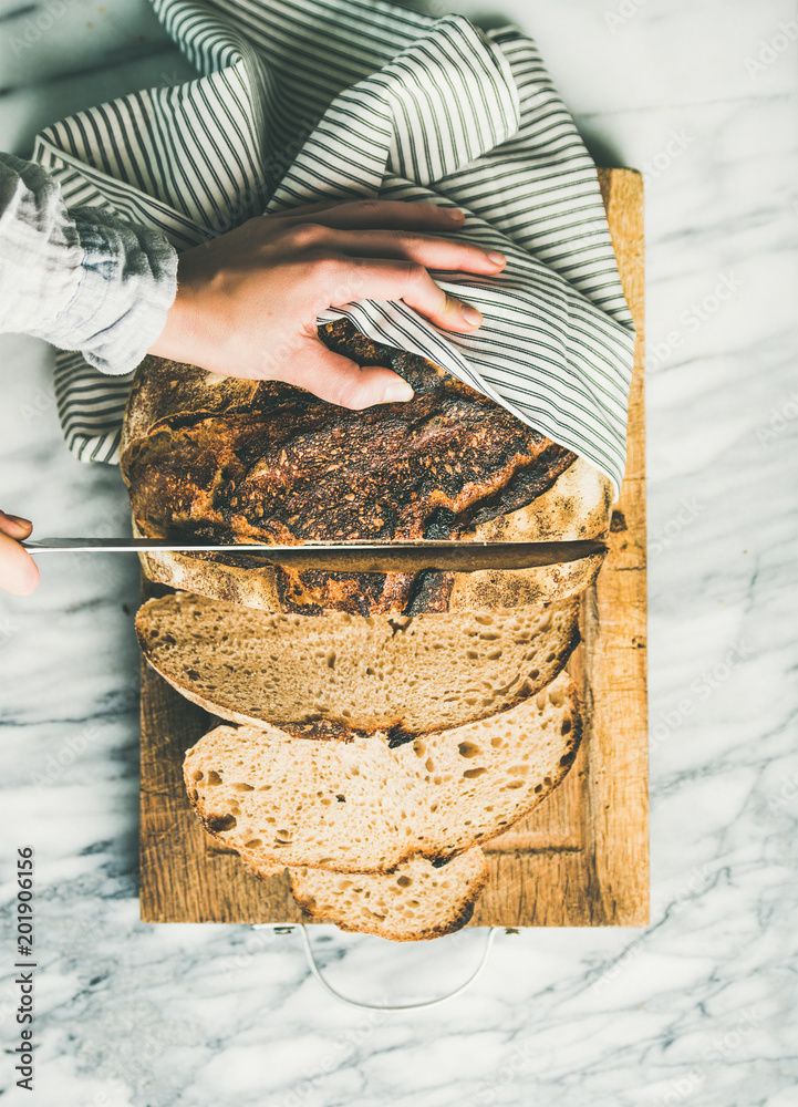 Flat-lay of woman's hands cutting freshly baked sourdough bread with knife into pieces on rustic wooden chopping board over light grey marble background, top view, vertical composition