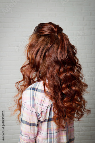 Hairstyle long curls on the red-haired girl turning head to the left on a light background.Professional female hairdress.