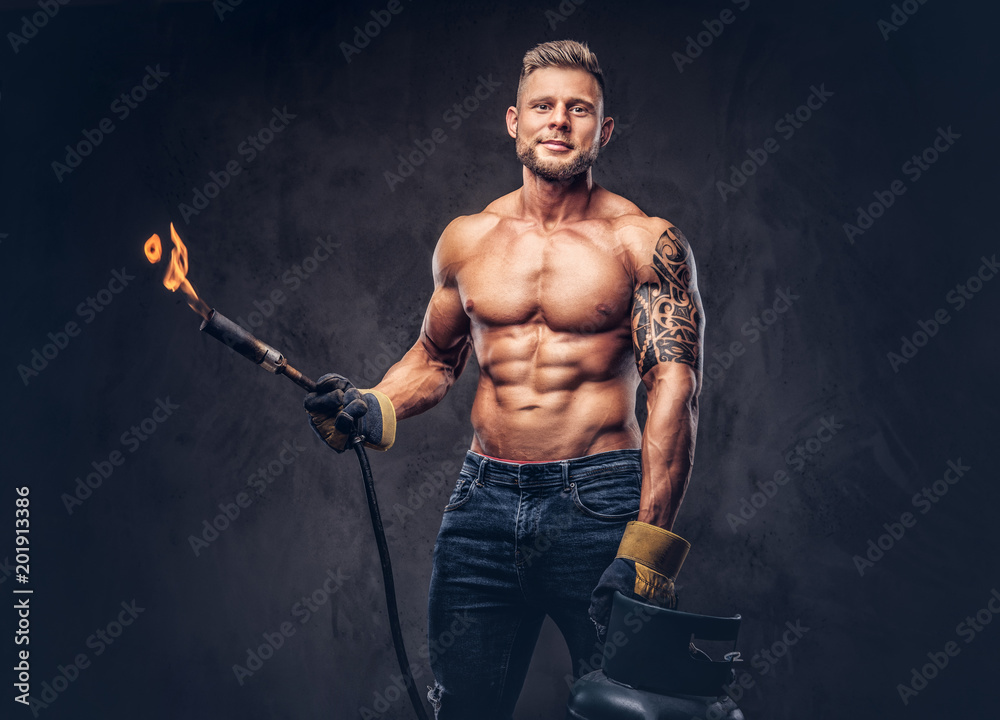 Brutal tattoed male welder with a stylish haircut and beard, with muscular body, dressed in only jeans, holds propane tank and a burning burner, standing in a studio, looking at a camera.