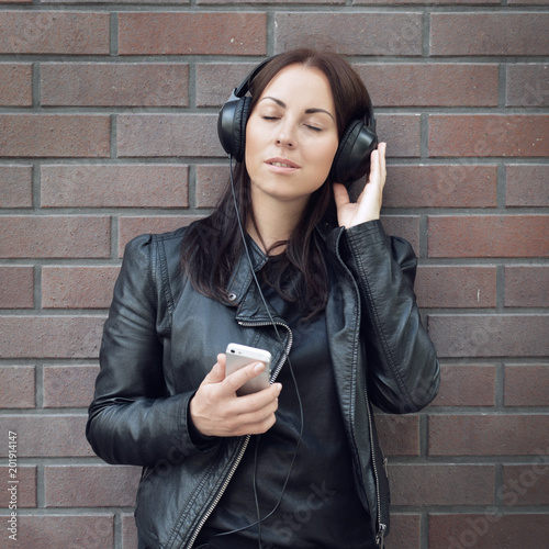A beautiful girl in a black leather jacket listens to music from external headphones from her phone. Brick wall. On the street. Complicating with music. New album.