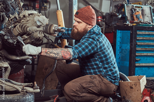 A brutal tattooed bearded mechanic specialist repairs the car engine which is raised on the hydraulic lift in the garage. Service station. © Fxquadro