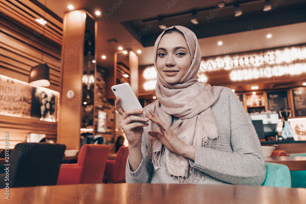 beautiful Arab girl with headscarf sitting in cafe, holding smartphone and looking at camera