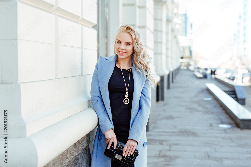 human emotion. Urban style. Portrait of a cute young blonde in a fashionable blue coat. The girl posing against the backdrop of urban architecture. human emotion. Urban style. Portrait of a cute young blonde in a fashionable blue coat. The girl posing against the backdrop of urban architecture.
