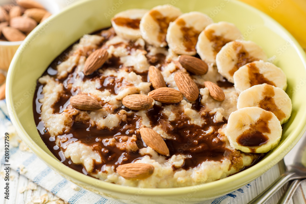 Oatmeal porridge with banana, almonds and chocolate syrup on white wooden background