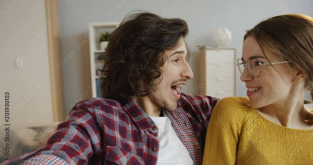 Close up of the young lovely smiling couple sitting on the gray couch and making nice selfies. Inside Close up of the young lovely smiling couple sitting on the gray couch and making nice selfies. Inside