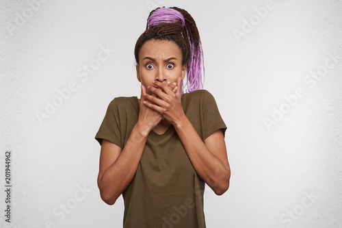 Indoor shot of shocked and amazed mixed race girl, looks directly into camera, keeps her hands on mouth. Facial expression. Isolated photo