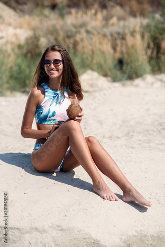 An attractive girl with fresh coconut relaxing on the beach. Summer vacation concept. Fashionable girl at sunglasses drinking cocktail on the beach. Young happy woman with healthy tanned body.
