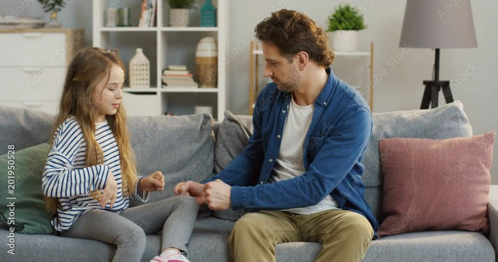Little blonde pretty girl sitting on the couch with her father and they playing a hands game. At home. Inside
