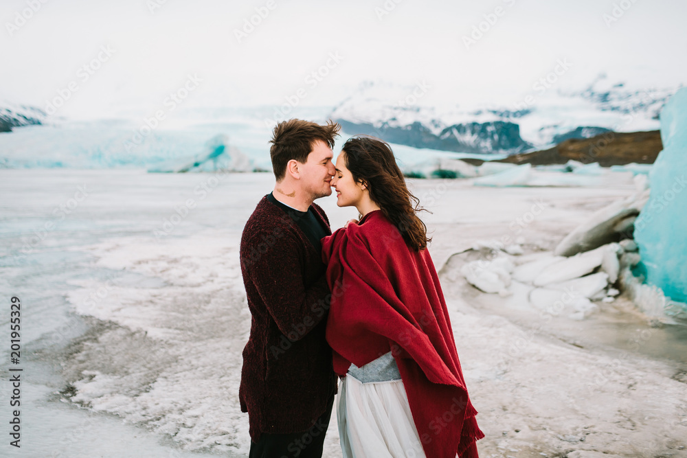 Groom tenderly kissing her beautiful bride in nose. Cheerful newlyweds is hugging in glacier lagoon, Iceland. . Newlyweds