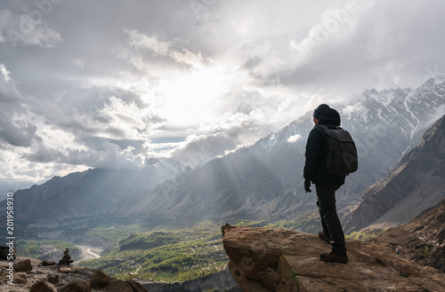 a man with backpack standing on mountain peak and bright sunlight through cloudy sky. Travel lifestyle and achievement success concept