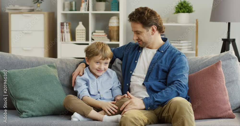 Young handsome Caucasian father sitting on the sofa in the living room and talking with his small cute son who sitting next to him. Indoors Young handsome Caucasian father sitting on the sofa in the living room and talking with his small cute son who sitting next to him. Indoors