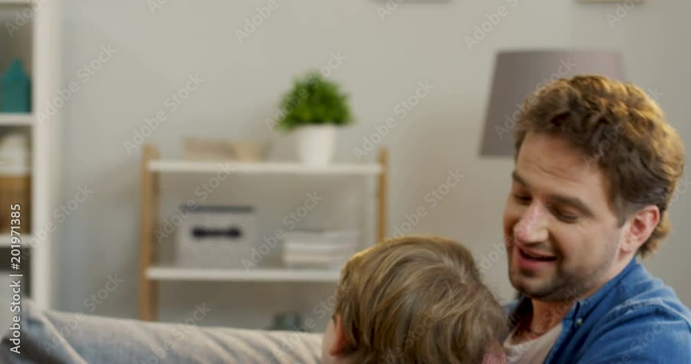 Close up of the Caucasian cheerful father and son having fun on the couch in the living room, fighting with pillows, tickling and laughing. Indoors Close up of the Caucasian cheerful father and son having fun on the couch in the living room, fighting with pillows, tickling and laughing. Indoors