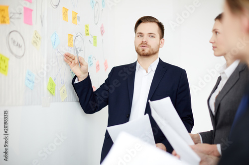 Businessman planning work with colleagues. He is standing and pointing at whiteboard