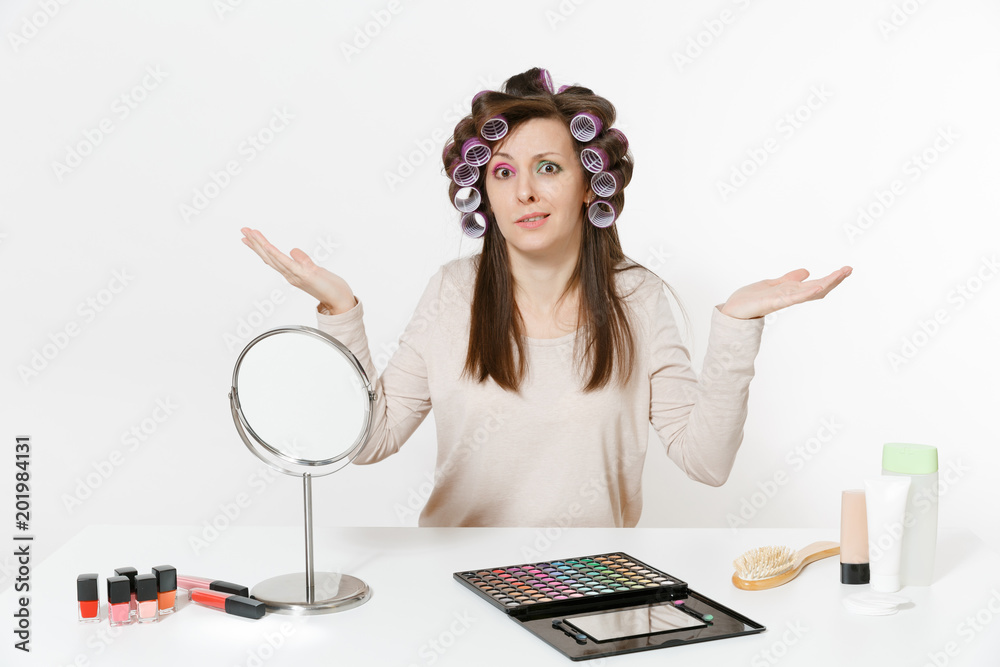 Irritated woman with curlers spreading hands, sitting at table applying makeup with set facial decorative cosmetics isolated on white background. Beauty female fashion lifestyle concept. Copy space.