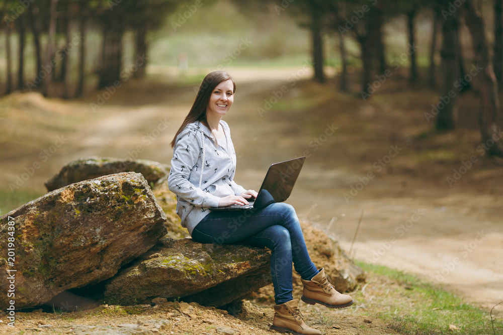 Young smiling successful smart business woman or student in casual clothes sitting on stone using laptop in city park or forest working outdoors on green blurred background. Mobile Office concept. Young smiling successful smart business woman or student in casual clothes sitting on stone using laptop in city park or forest working outdoors on green blurred background. Mobile Office concept.