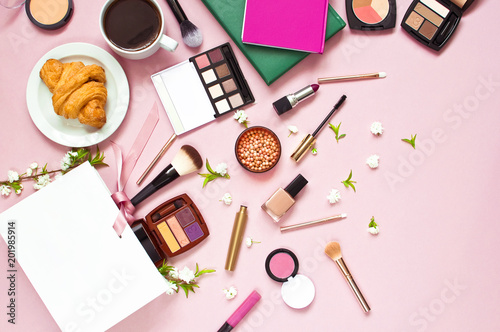 Various cosmetic products for makeup and white spring flowers in a white gift bag, notepad, cup of coffee, croissant on a pink pastel background top view with copy space