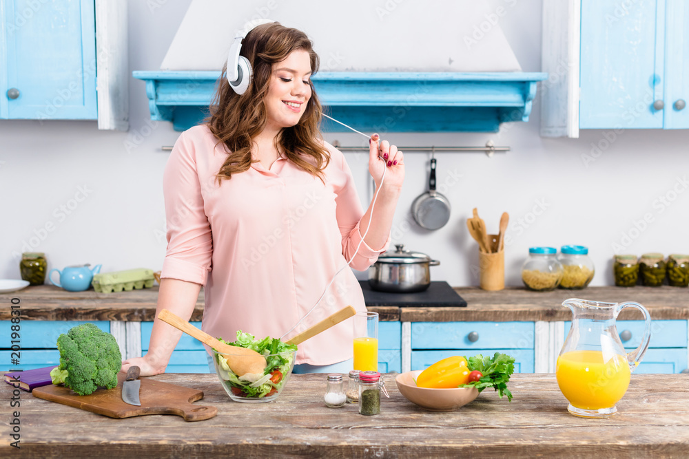 cheerful overweight woman listening music in headphones at table with fresh vegetables in kitchen at home