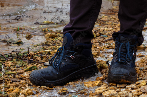 A lonely man in dark jeans and heavy road boots on lacing strolls in the cold season stands on the river bank. On the shore, small stones, ice, thickets of reeds and shrubs.