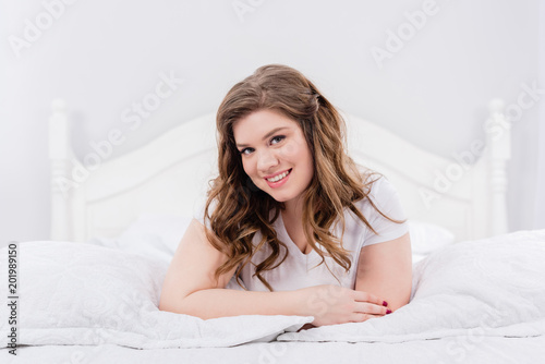 portrait of smiling young woman lying on bed and looking at camera at home