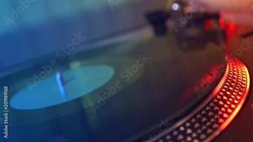 Vinyl player. A fragment of a rotating vinyl plate, a woman's hand puts vinyl and lowers the stylus with a close-up needle. Macro. Foreground. Blurred against the background of red and blue lights