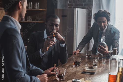 three multiethnic men in suits smoking cigars and drinking alcohol together