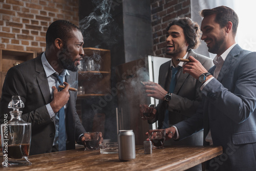 smiling multiethnic male friends in suits smoking cigars, drinking whiskey and talking photo