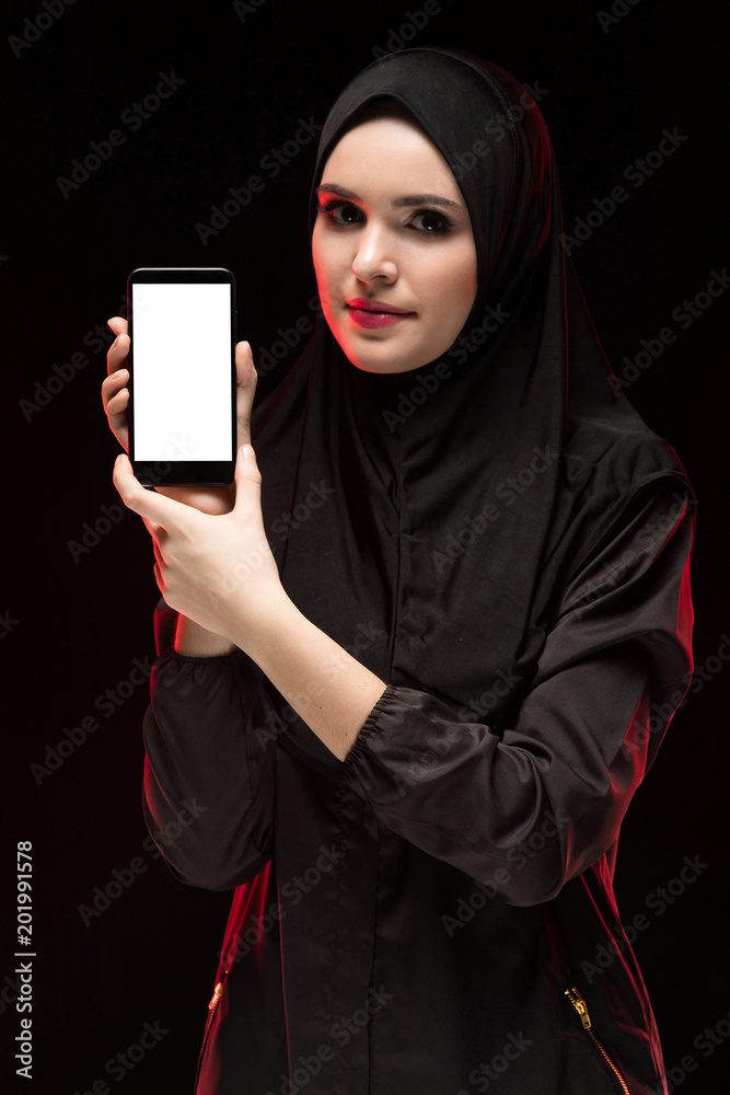 Portrait of beautiful smart young muslim woman wearing black hijab advertising mobile phone in her hands as education concept on black background Portrait of beautiful smart young muslim woman wearing black hijab advertising mobile phone in her hands as education concept on black background