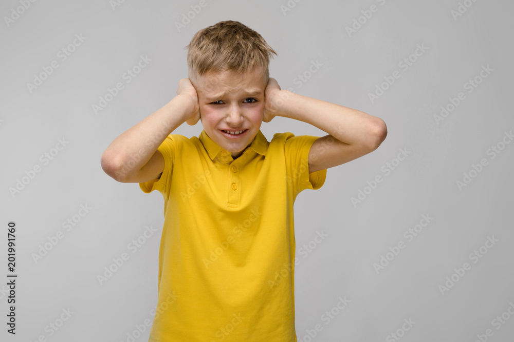 Portrait of blonde caucasian sad little boy in yellow t-shirt closing his ears with hands on gray background Portrait of blonde caucasian sad little boy in yellow t-shirt closing his ears with hands on gray background