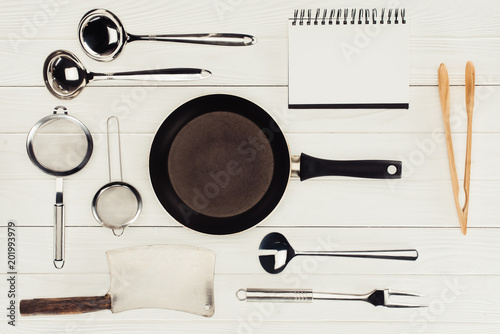 top view of blank textbook, frying pan, butcher axe and kitchen utensils on white wooden table