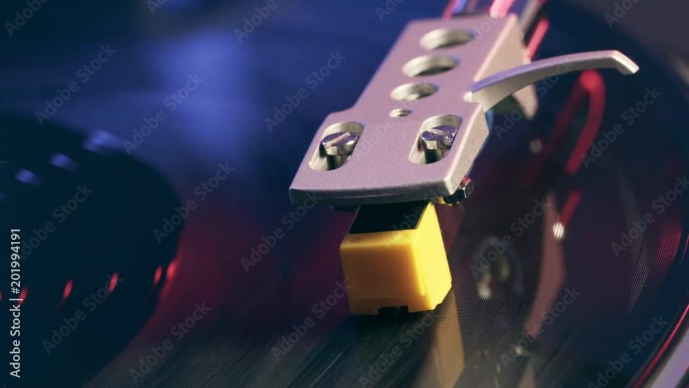 The player vinyl turntable close-up on a background of red and blue lights. Fragment of a rotating black vinyl record player. Rotating plate and stylus with a needle turntable close-up