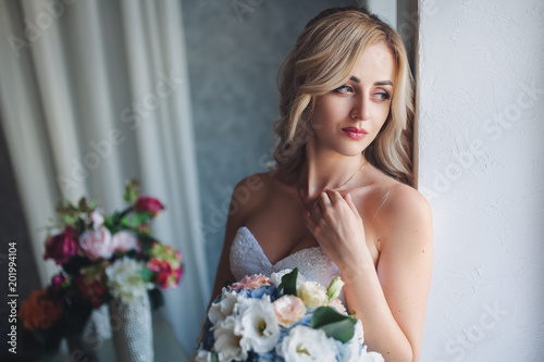 Portrait of a beautiful bride with curly hair in a gray studio with flowers. A sweet bride gently stands near a window in a modern studio with a chic decor, passing her hand over her body.