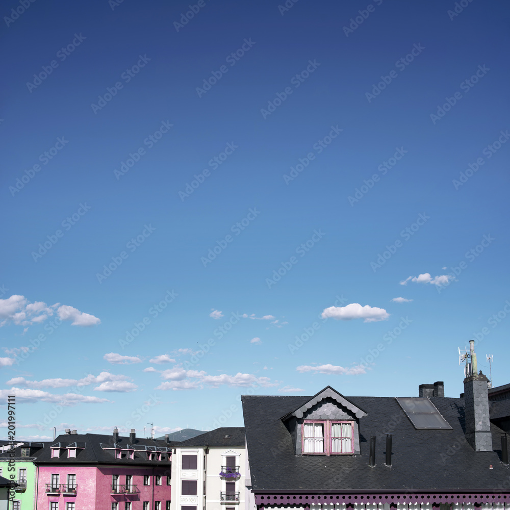 Minimal urban picture of some candy-colored buildings’ rooftops from a little village. Day scene with a wide clear and blue sky with some clouds.