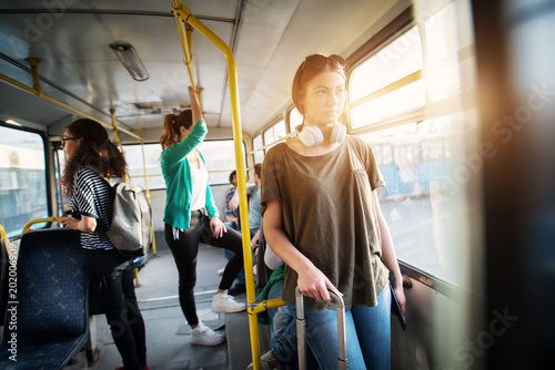Young charming woman with headset around her neck traveling by bus and holding her luggage while leaning against the bus bars.