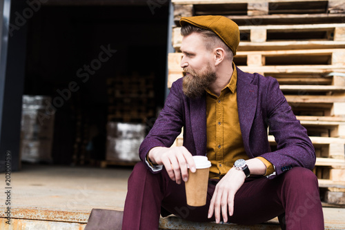 Stylish bearded businessman sitting on street with paper cup in hands