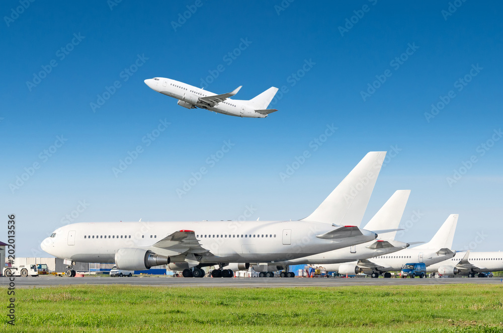 Obraz premium Rząd samolotów pasażerskich, samolot zaparkowany na służbie przed odlotem na lotnisku, inny samolot odepchnął hol. Jeden startuje z pasa startowego na niebieskim niebie.