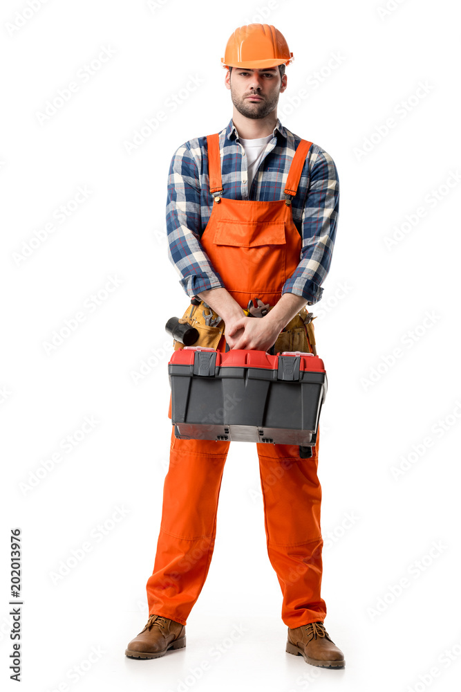 Confident handyman in orange overall and hard hat holding tool box isolated on white Confident handyman in orange overall and hard hat holding tool box isolated on white