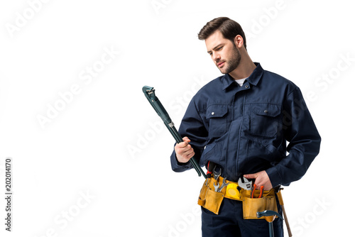 Bearded sanitary engineer wearing uniform with tool belt and looking at wrench isolated on white