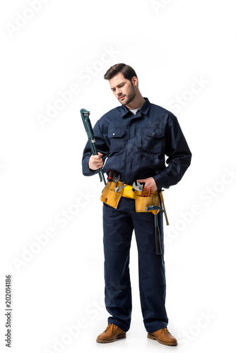 Young handyman wearing uniform with tool belt and looking at wrench isolated on white