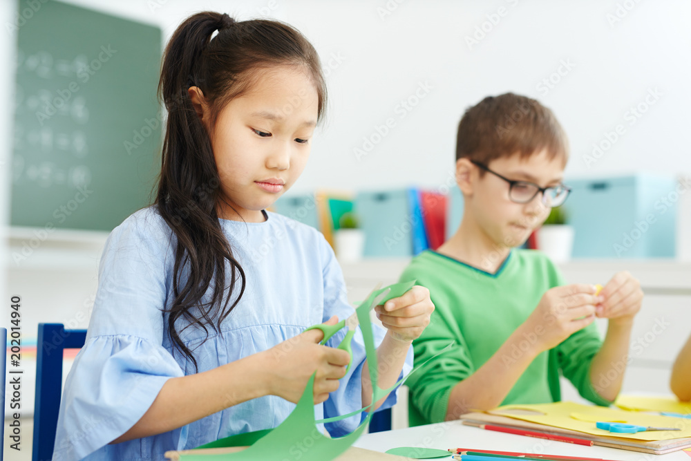 Little Asian girl and her Caucasian classmate cutting colored paper with scissors while making origami on art lesson at school Little Asian girl and her Caucasian classmate cutting colored paper with scissors while making origami on art lesson at school