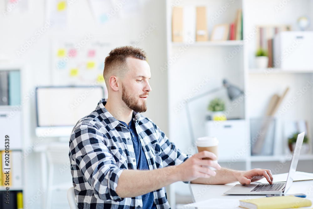 Designer using laptop for work and drinking coffee at office