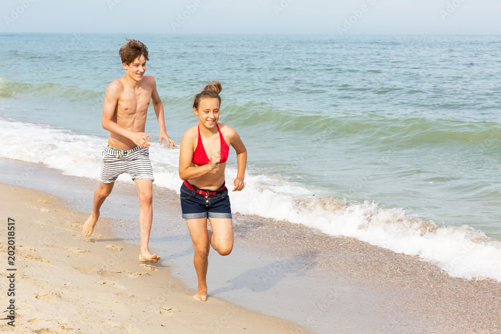 Two teenagers: a girl and a boy with blond hair, dressed in a swimsuit run, laugh and play in the sea beach.